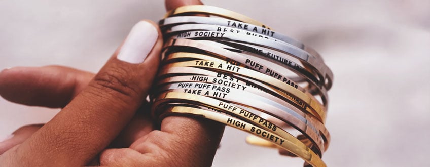 Puff Puff Pass Bracelets Een hand die een stapel gouden en zilveren armbanden vasthoudt met verschillende ingegraveerde zinnen.