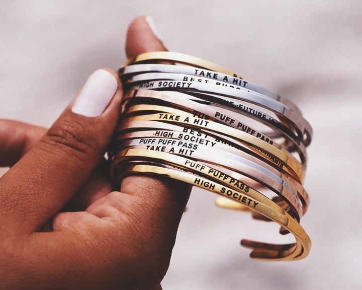 Puff Puff Pass Bracelets Een hand die een stapel gouden en zilveren armbanden vasthoudt met verschillende ingegraveerde zinnen.